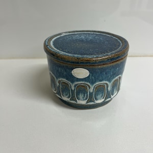May include: A small, round ceramic container with a lid. The container is a deep blue with a brown rim and a repeating white and brown oval pattern around the base. A small, silver sticker is affixed to the side.