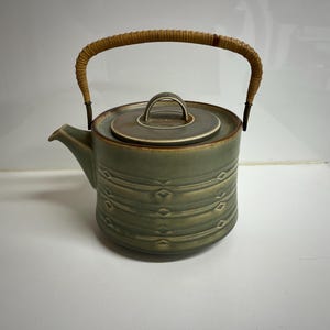 Puede incluir: Una tetera de cerámica verde con un asa de madera trenzada y una tapa. La tetera tiene un patrón geométrico.