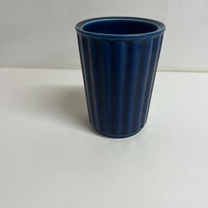 Può includere: Un bicchiere in ceramica blu navy con dettagli a coste verticali. La tazza cilindrica ha una parte superiore leggermente svasata e un interno blu scuro. Il design è semplice ed elegante, adatto a varie bevande.