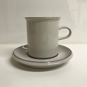 Könnte beinhalten: Eine weiße Kaffeetasse aus Keramik mit braunem Rand und einer passenden Untertasse. Die Tasse hat ein schlichtes Design und einen abgerundeten Henkel.
