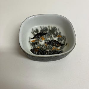 Può includere: Un piccolo piatto quadrato in ceramica con interno bianco ed esterno grigio scuro. L'interno presenta un motivo floreale nero, arancione e grigio. Il piatto viene probabilmente utilizzato per servire piccole porzioni di cibo o come oggetto decorativo.