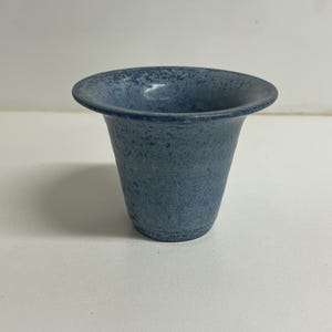 Puede incluir: Un pequeño jarrón de cerámica azul con un esmalte moteado. El jarrón tiene una abertura ancha y acampanada y una base estrecha.