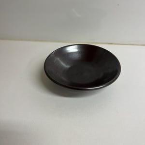 Puede incluir: Un cuenco pequeño, redondo y marrón oscuro con un acabado brillante. El cuenco está vacío y se encuentra sobre una superficie blanca. La forma del cuenco es simple y clásica, adecuada para servir porciones pequeñas.