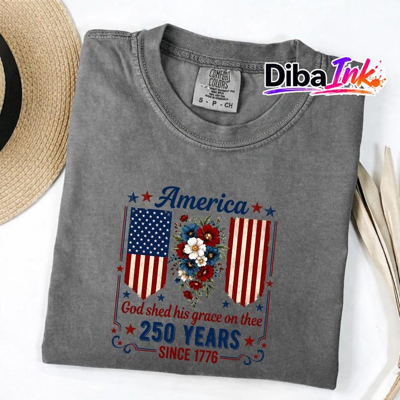 May include: Grey t-shirt with a patriotic design. The shirt displays the word "America" above a floral arrangement and two American flags. Below the flags, the text reads "God shed his grace on thee" and "250 YEARS SINCE 1776".