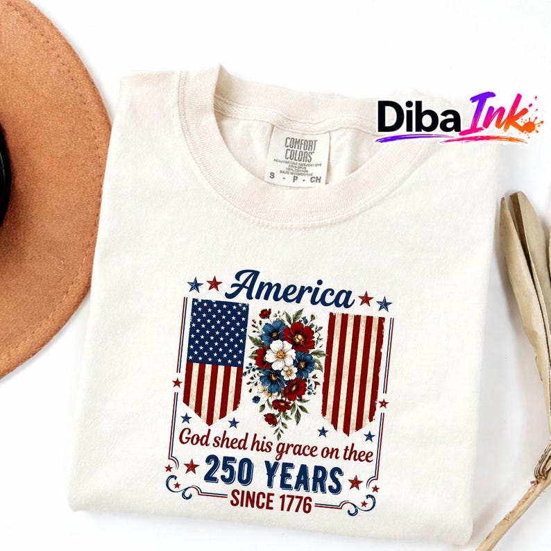 May include: Off-white t-shirt with a patriotic design. The design includes the American flag, floral elements, and the text "America," "God shed his grace on thee," and "250 YEARS SINCE 1776."