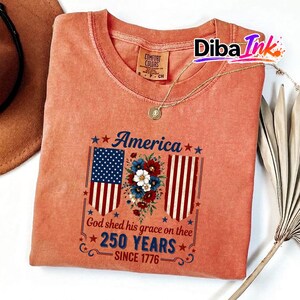 May include: A coral-coloured t-shirt with a patriotic design. The graphic includes the word "America" above a floral arrangement and American flag imagery. The text "God shed his grace on thee 250 YEARS SINCE 1776" is also displayed.