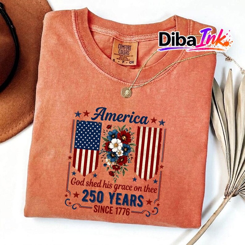 May include: A coral-coloured t-shirt with a patriotic design. The graphic includes the word "America" above a floral arrangement and American flag imagery. The text "God shed his grace on thee 250 YEARS SINCE 1776" is also displayed.