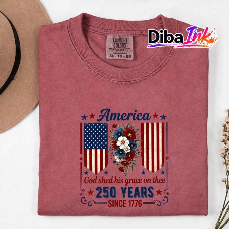 May include: A maroon t-shirt featuring the word "America" and the text "God shed his grace on thee 250 YEARS SINCE 1776". The design incorporates American flags and floral arrangements, suitable for casual wear.