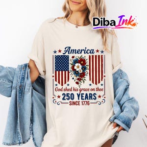 May include: A cream-coloured t-shirt with a patriotic design. The graphic features the word "America" above a stylized American flag and floral arrangement. Below, the text reads "God shed his grace on thee 250 YEARS SINCE 1776."