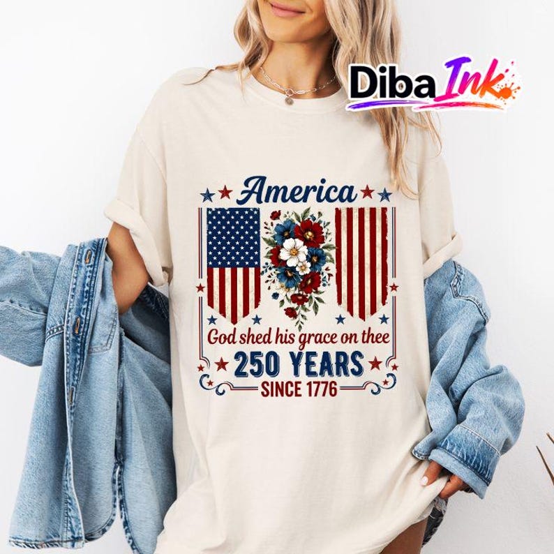 May include: A cream-coloured t-shirt with a patriotic design. The graphic features the word "America" above a stylized American flag and floral arrangement. Below, the text reads "God shed his grace on thee 250 YEARS SINCE 1776."