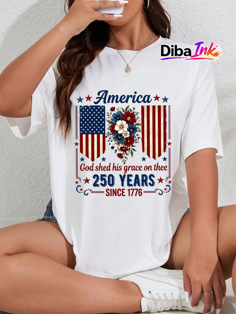May include: White t-shirt with a patriotic design. The design features the word "America" above a floral arrangement and the American flag. Below the flag are the words "God shed his grace on thee" and "250 YEARS SINCE 1776."