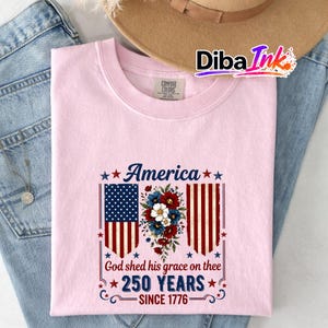 May include: Pale pink t-shirt featuring the word "America" and a floral design. The design includes the American flag and the text "God shed his grace on thee 250 YEARS SINCE 1776."