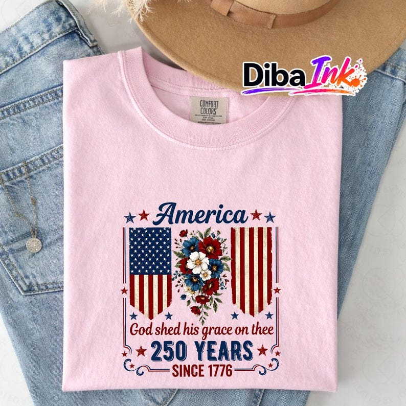 May include: Pale pink t-shirt featuring the word "America" and a floral design. The design includes the American flag and the text "God shed his grace on thee 250 YEARS SINCE 1776."