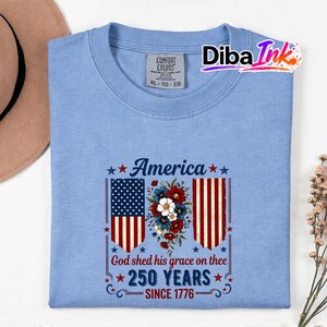 May include: A pale blue t-shirt featuring a patriotic design. The design includes the word "America" above a floral arrangement and the American flag. The text "God shed his grace on thee 250 YEARS SINCE 1776" is also featured.