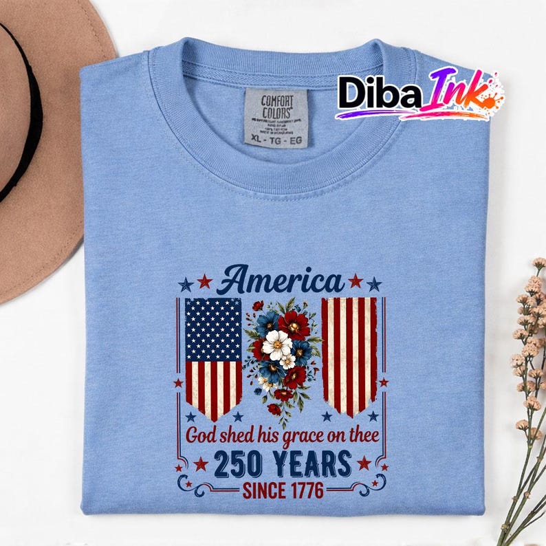 May include: A pale blue t-shirt featuring a patriotic design. The design includes the word "America" above a floral arrangement and the American flag. The text "God shed his grace on thee 250 YEARS SINCE 1776" is also featured.