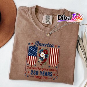 May include: A brown Comfort Colors t-shirt featuring a patriotic design. The graphic includes the American flag, floral elements, and the text "America", "God shed his grace on thee", and "250 YEARS SINCE 1776". A silver necklace is draped on the shirt.