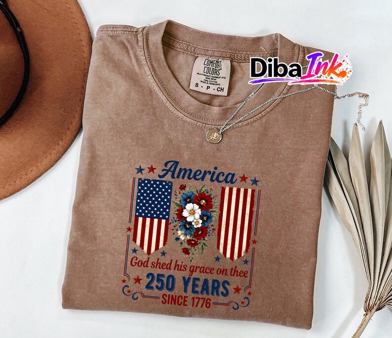 May include: A brown Comfort Colors t-shirt featuring a patriotic design. The graphic includes the American flag, floral elements, and the text "America", "God shed his grace on thee", and "250 YEARS SINCE 1776". A silver necklace is draped on the shirt.