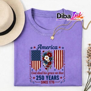 May include: A lilac t-shirt featuring the word "America" and the text "God shed his grace on thee 250 YEARS SINCE 1776". The design incorporates American flags and floral motifs. A straw hat and a gold necklace are also visible.