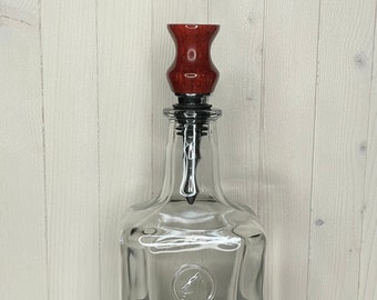 Puede incluir: Una botella de vidrio transparente con un tapón de botella de madera roja. La botella tiene una superficie texturizada y un fondo redondo.