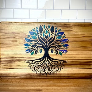 May include: Wooden serving tray with a tree of life design. The tray is rectangular with integrated handles and a natural wood grain finish. The tree design features blue and teal leaves and black branches.