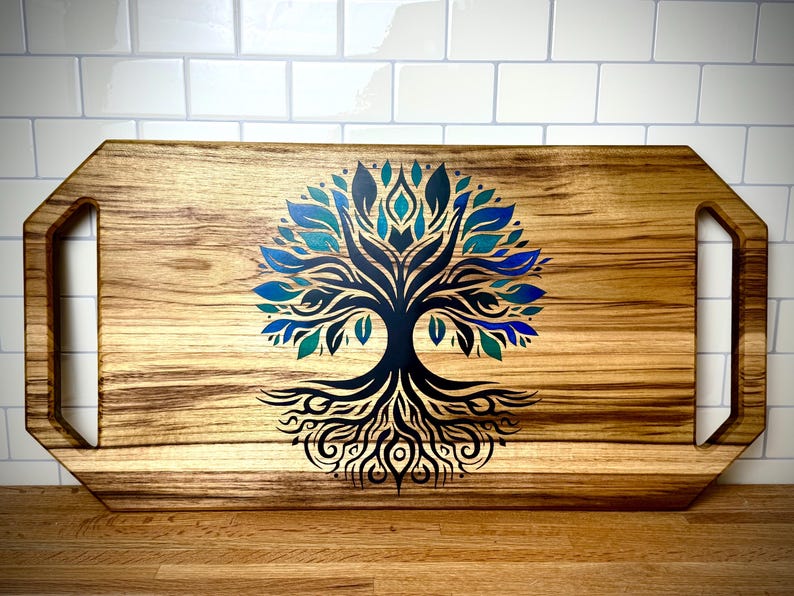 May include: Wooden serving tray with a tree of life design. The tray is rectangular with integrated handles and a natural wood grain finish. The tree design features blue and teal leaves and black branches.