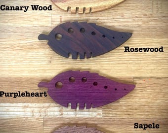 Puede incluir: Cuatro herramientas de madera con forma de hoja con diferentes patrones de veta de madera. Las herramientas están etiquetadas como "Canary Wood", "Rosewood", "Purpleheart" y "Sapele".