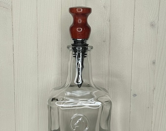 Puede incluir: Una botella de vidrio transparente con un tapón de madera. El tapón es de color rojo oscuro y tiene una parte superior lisa y redondeada. La botella está vacía y tiene una superficie texturizada.