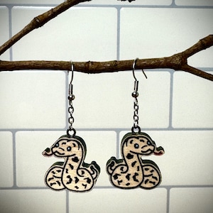 May include: A pair of wooden snake earrings with black and brown markings. The earrings are hanging from silver chains.