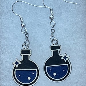 May include: A pair of silver earrings with a black and blue potion bottle charm. Each bottle has a white star on the side and a blue liquid inside.