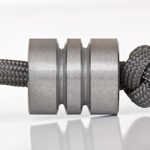 May include: A gray metal bead with three grooves, attached to a gray braided cord. The cord is knotted at the end. The bead has a matte finish and is centered in the frame.