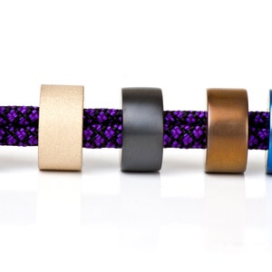 May include: Five different colored metal beads strung on a purple and black cord. The beads are silver, gold, black, copper, and blue.