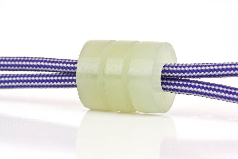 May include: A close-up of a white cord lock with a purple and white striped cord threaded through it.