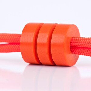 May include: A close-up of a bright orange cord stopper with three stacked rings. The cord stopper is made of a smooth, plastic material and is threaded through a red cord.