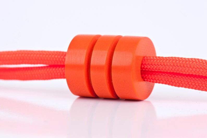 May include: A close-up of a bright orange cord stopper with three stacked rings. The cord stopper is made of a smooth, plastic material and is threaded through a red cord.