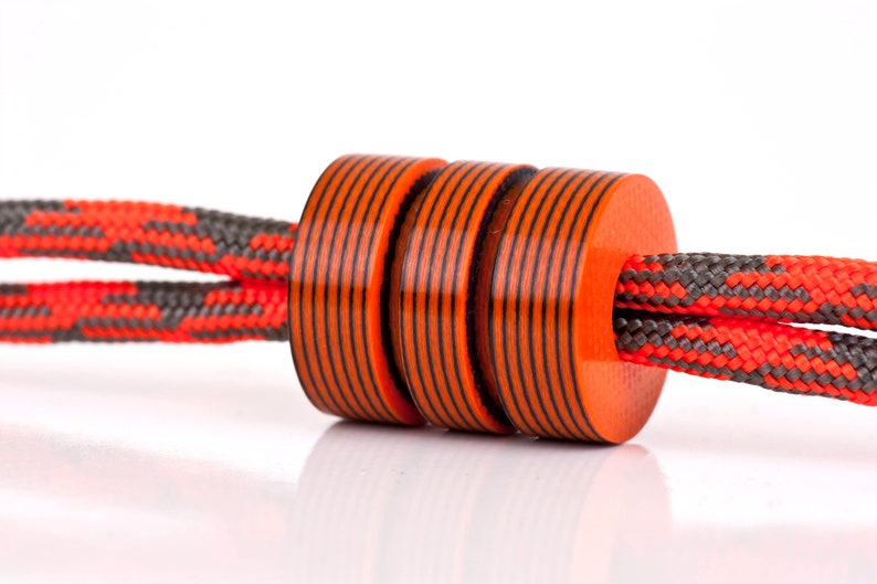 May include: Three orange and black striped beads are strung on a red and gray rope. The beads are cylindrical and have a glossy finish.