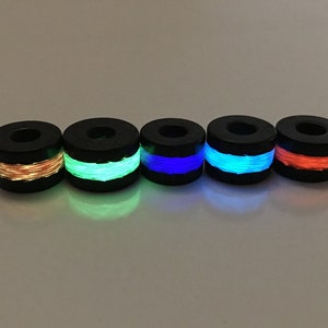 May include: Five black cylindrical beads with a single band of glow-in-the-dark filament. The filament colors are orange, green, blue, light blue, and red.
