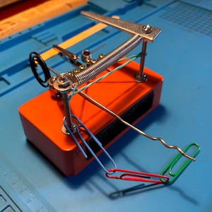 May include: A rectangular orange base with a mechanical assembly featuring a horizontal metal spring, two vertical threaded rods with nuts, a flat metal arm, and a wavy wire extension. A chain of blue, red, and green paper clips is attached.