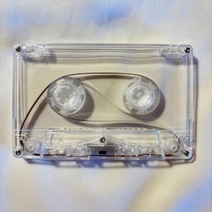 Op de afbeelding: Een transparante audiocassette met zichtbare spoelen en magnetische tape. De cassette is rechthoekig met afgeronde hoeken en kleine schroeven. Een vintage item voor audio-opname.