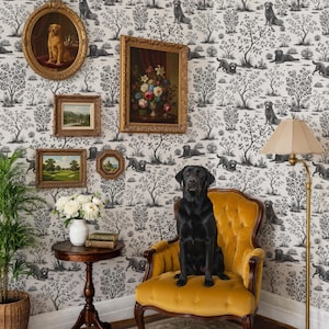 May include: A black Labrador sits on a yellow velvet armchair in a room with dog-themed wallpaper and framed artwork. A small side table holds a vase of white flowers and books. A floor lamp provides soft lighting.
