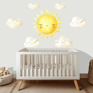 May include: A white crib with a wooden frame and legs, set against a wall decorated with a watercolor-style sun and cloud decals. The sun has a smiling face. A woven basket and a rug are in the foreground.