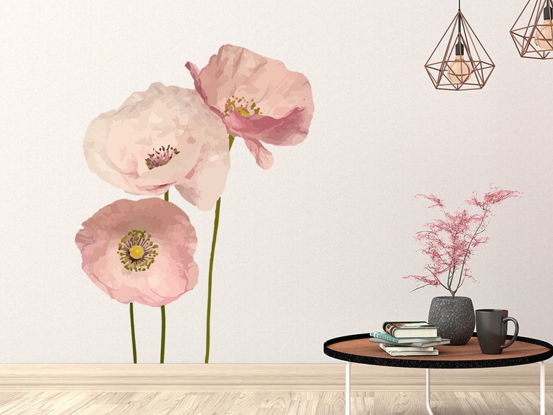 May include: Three pink poppy flowers with yellow centers on a white wall. The flowers are in a watercolor style and are arranged in a vertical line. There is a small table with a potted plant and books on it in the bottom right corner of the image.