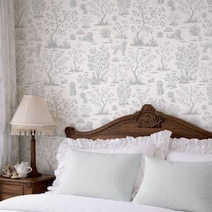 May include: A bedroom scene featuring a wooden headboard, white bedding, and light blue pillows. The wallpaper has a repeating pattern of dogs and trees in a soft, muted color palette. A lamp with a fringed shade sits on a wooden nightstand.