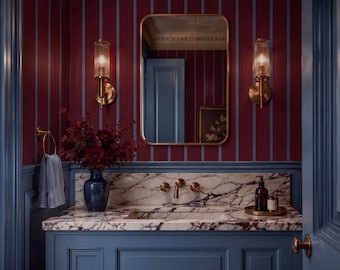 May include: A bathroom vanity featuring a white marble countertop with deep red veining, a blue cabinet base, and a brass faucet. Above, a rectangular mirror is flanked by two ribbed glass wall sconces against dark red and blue striped wallpaper.
