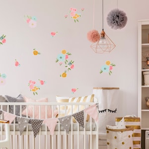 May include: A white crib with a pink and gray patterned crib bumper and a pink and gray patterned bunting. The crib is in a nursery with a white bookshelf, a white chair, and a pink and gray pom-pom garland hanging from the ceiling.