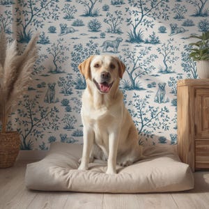 以下が含まれることがあります： ベージュの犬用ベッドに座る、明るい黄色のラブラドールレトリバー。犬は、犬と木のイラストが描かれた青と白のトワルドジュイの壁紙の前にいます。左側には、乾燥した植物が入ったバスケットがあります。