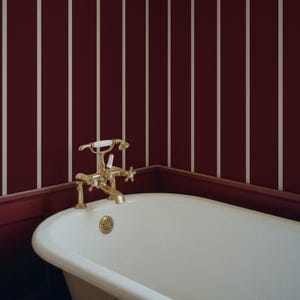 Puede incluir: Interior de baño con bañera blanca y grifería dorada. Las paredes son de color burdeos oscuro con rayas blancas verticales. La grifería tiene manijas blancas y un diseño clásico.