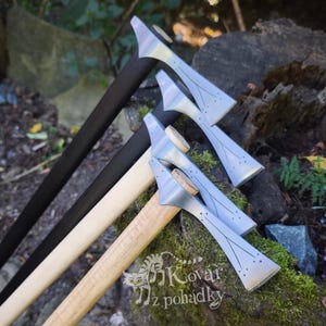 May include: Four axes with silver-colored metal heads and wooden handles. Two handles are dark brown and two are light tan. The metal heads feature a geometric engraved design. A logo with a stylized cat and the text Kovar z pohádky is visible.