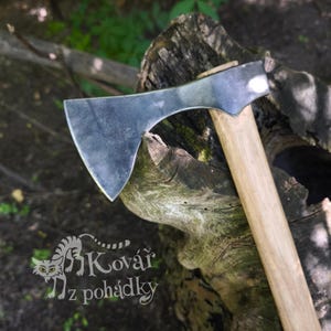 May include: A hand-forged steel axe head with a wide, curved blade attached to a smooth, light-colored wooden handle. The metal surface shows a dark, textured finish. A logo featuring a stylized cat and the text Kovář z pohádky is visible.