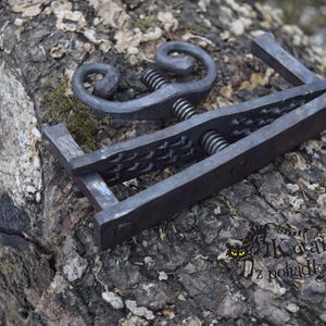 Könnte beinhalten: Eine schwarze Metallklemme mit einem dekorativen Schnörkeldesign und einer Gewindeschraube. Die Klemme ist aus Schmiedeeisen gefertigt und hat eine strukturierte Oberfläche.