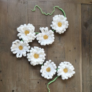 May include: A crocheted daisy garland featuring seven white flowers with yellow centers, connected by a green crocheted vine. The garland is displayed on a wooden surface.